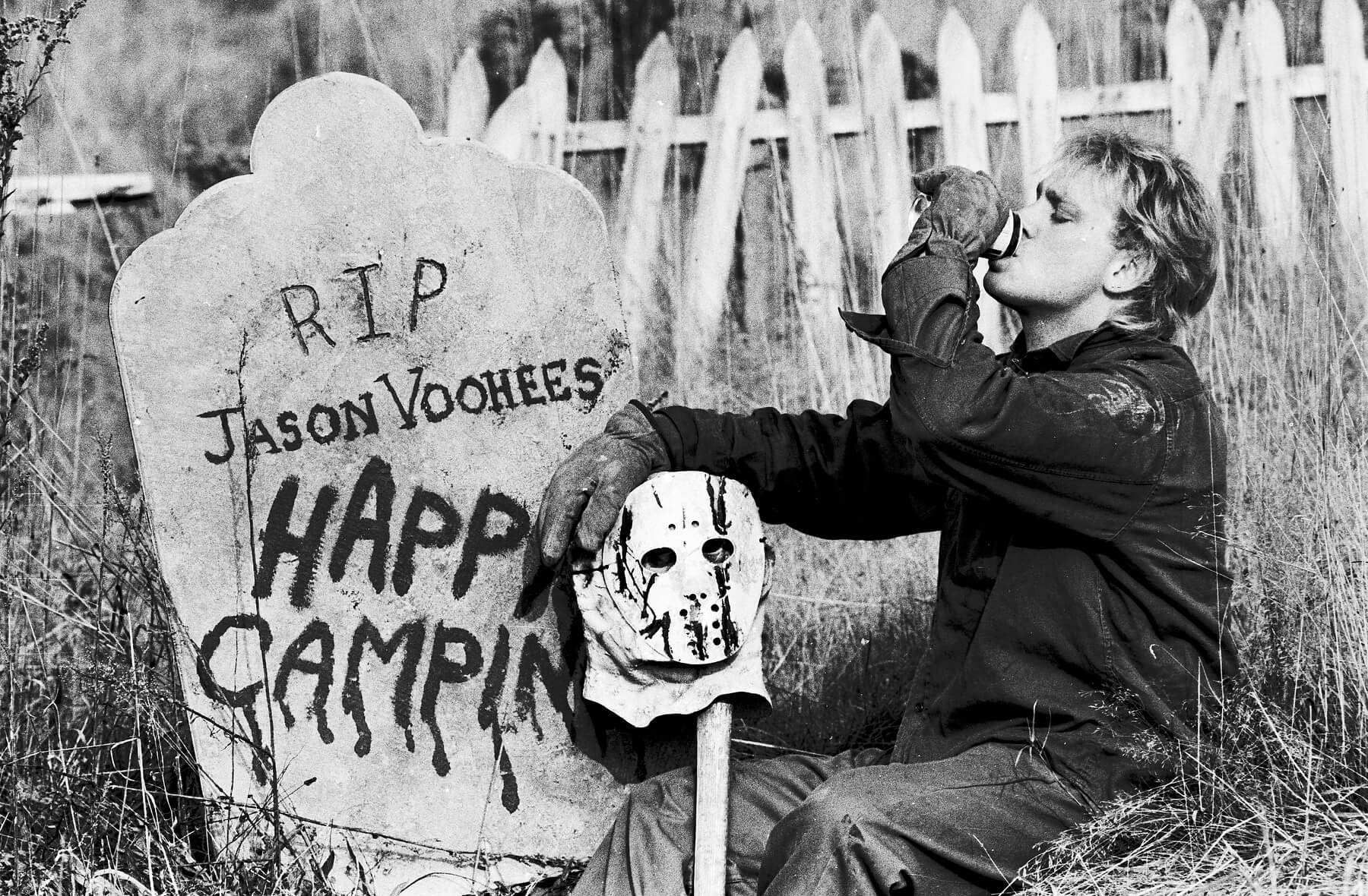 A halloween haunt performer dressed as Jason Voorhees takes a break, drinking a soda while sitting among tombstones.