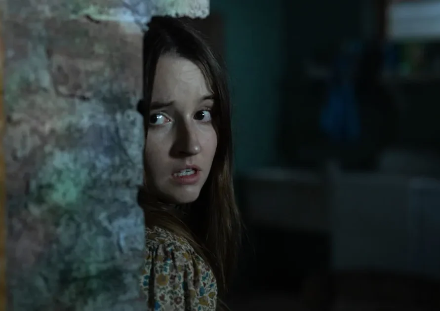 No One Will Save You (2023) A Twenty-Something Brynn Hides From Aliens, peaking around the edge of a stone farmhouse wall