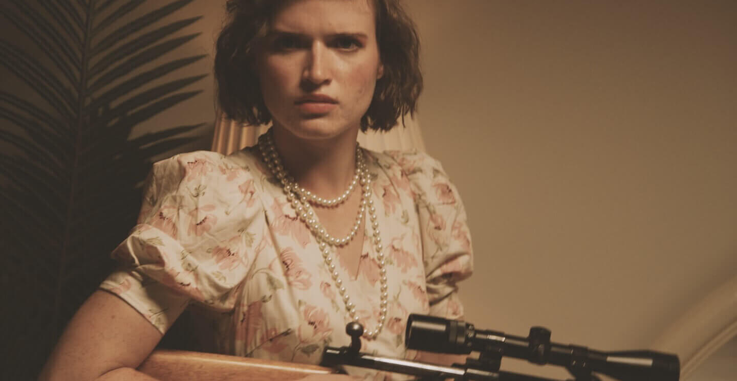 A mad teenage girl wearing pearls and a flowery dress looks directly into the camera. She's holding a hunting rifle and she's eager to use it.