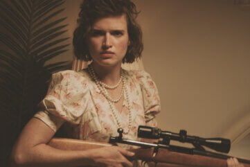 A mad teenage girl wearing pearls and a flowery dress looks directly into the camera. She's holding a hunting rifle and she's eager to use it.