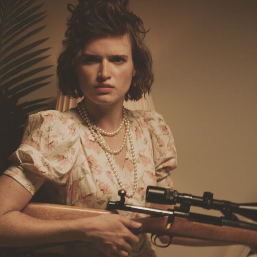 A mad teenage girl wearing pearls and a flowery dress looks directly into the camera. She's holding a hunting rifle and she's eager to use it.