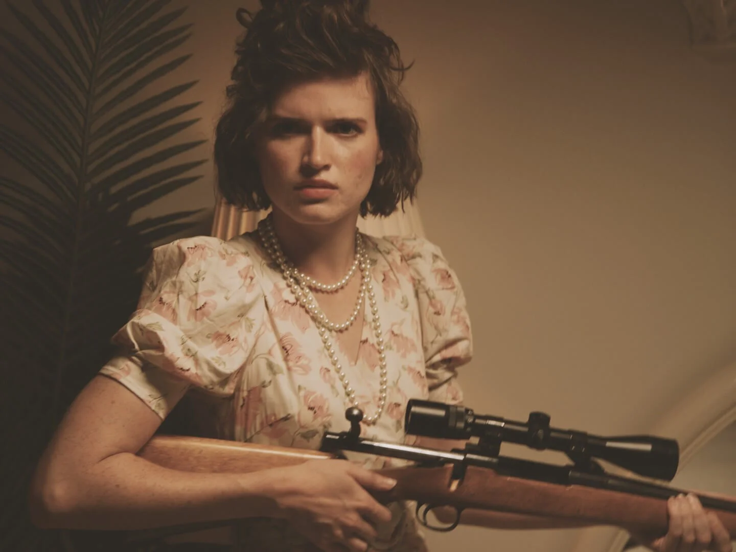 A mad teenage girl wearing pearls and a flowery dress looks directly into the camera. She's holding a hunting rifle and she's eager to use it.